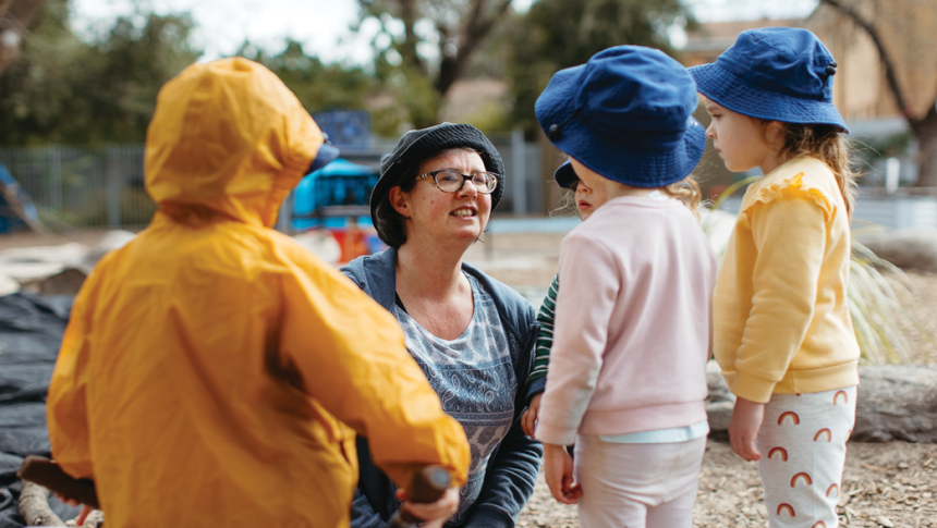 An early childhood educator outside with three children. The educator is kneeling down to speak to them at their level. 
