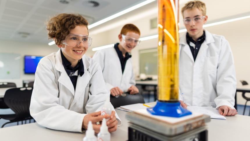 3 male students happily conducting a science experiment wearing lab cots and goggles.