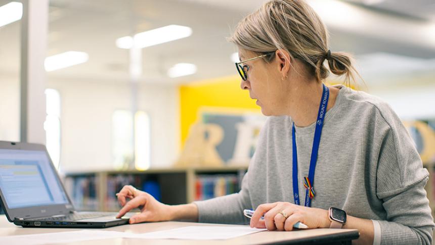 A teacher sitting at a table in a library using a laptop