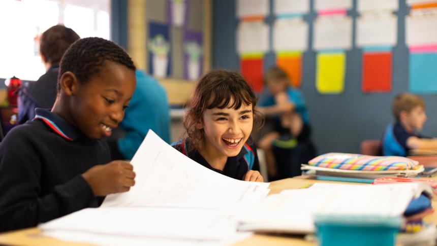 Two primary-school-aged children sit at a desk in a classroom. One is smiling and leafing through a workbook and the other is laughing.