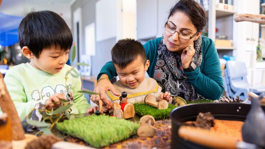 Two primary-school-aged children sit at a desk in a classroom. One is smiling and leafing through a workbook and the other is laughing.