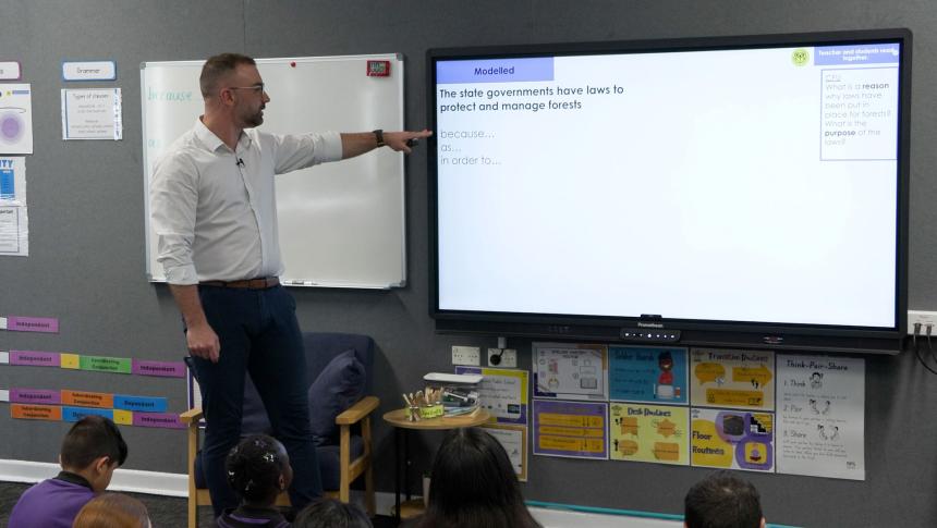 Teacher in front of class pointing at a grammar lesson on a large screen