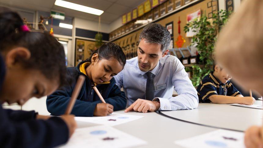 A teacher sitting at a low desk with several primary school students helping them with writing