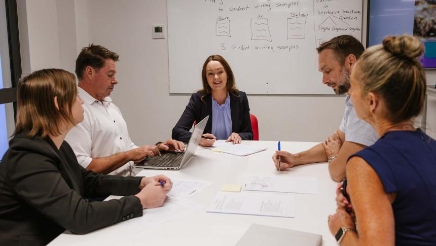 A group of educators having a discussion at a meeting room table with papers and a laptop