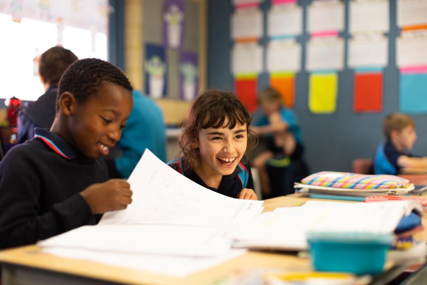 Two primary-school-aged children sit at a desk in a classroom. One is smiling and leafing through a workbook and the other is laughing.